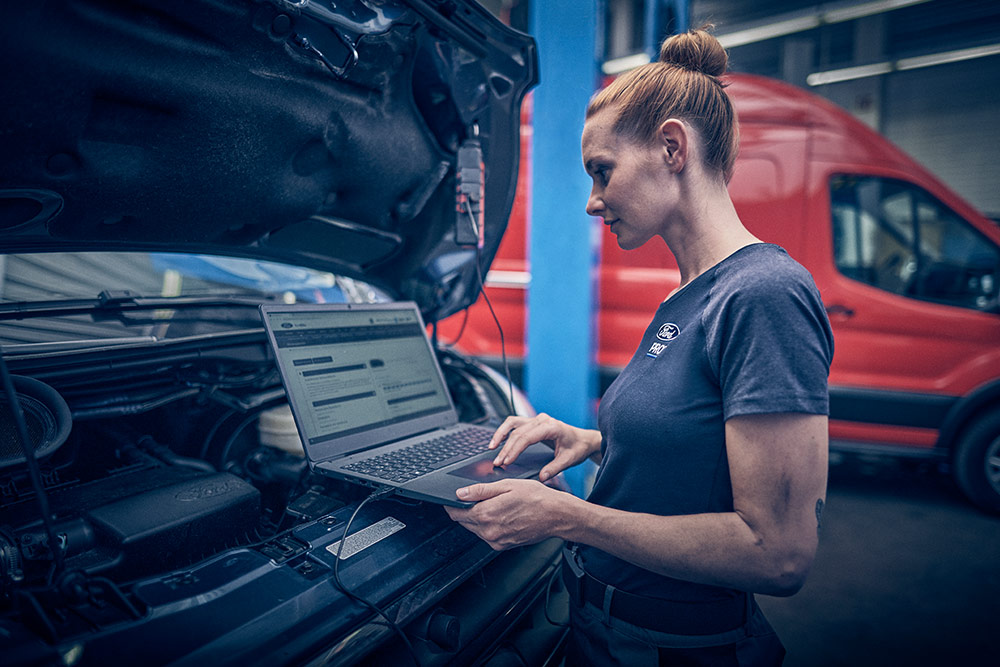 Een technicus diagnosticeert een Ford-voertuig met laptop, motorkap open, en een rode Ford Transit op de achtergrond.