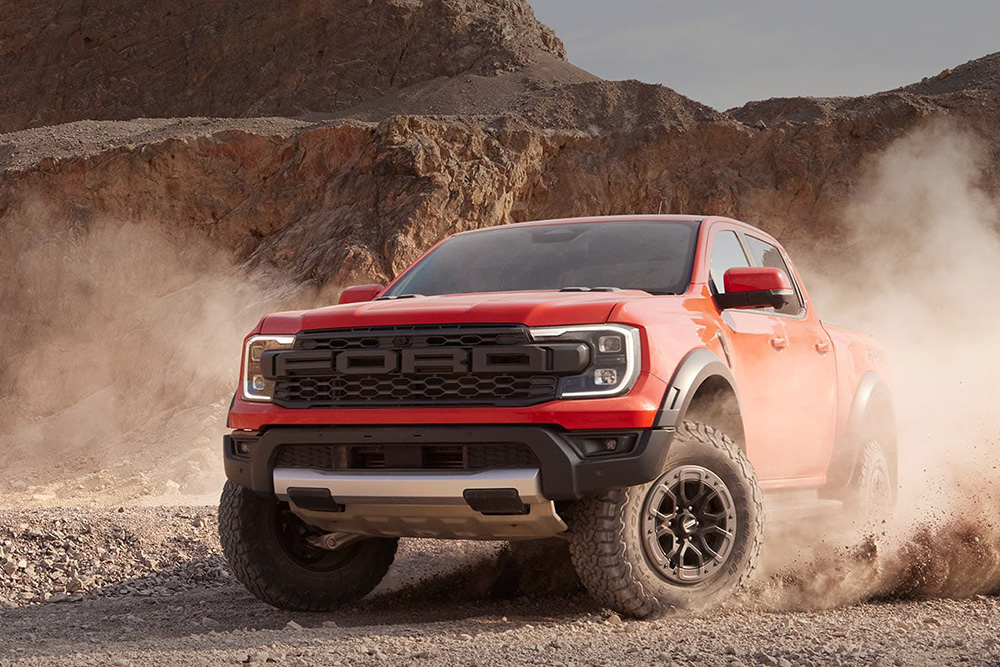Oranje Ford Ranger Raptor rijdt door het stof in een rotsachtig landschap. 