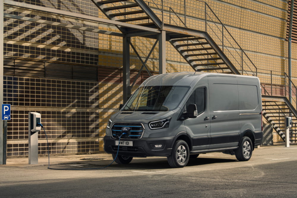 Een grijze elektrische Ford E-Transit bedrijfswagen die wordt opgeladen bij een laadpaal tegen de achtergrond van een modern industrieel gebouw met metalen trappen.