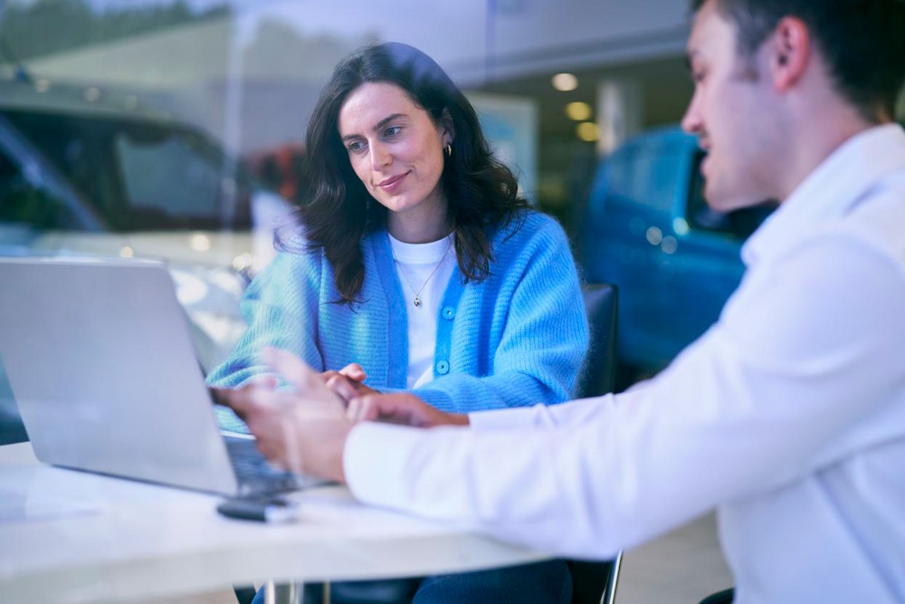 Twee personen in gesprek aan een tafel met een laptop, op de achtergrond is een Ford-voertuig zichtbaar.