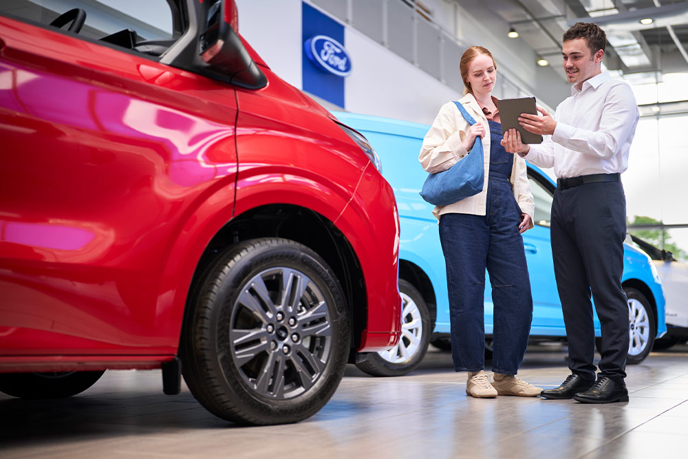 Twee personen in een Ford-showroom kijken naar een tablet, met een rode Ford-bestelwagen op de voorgrond en een blauwe Ford-bestelwagen erachter.