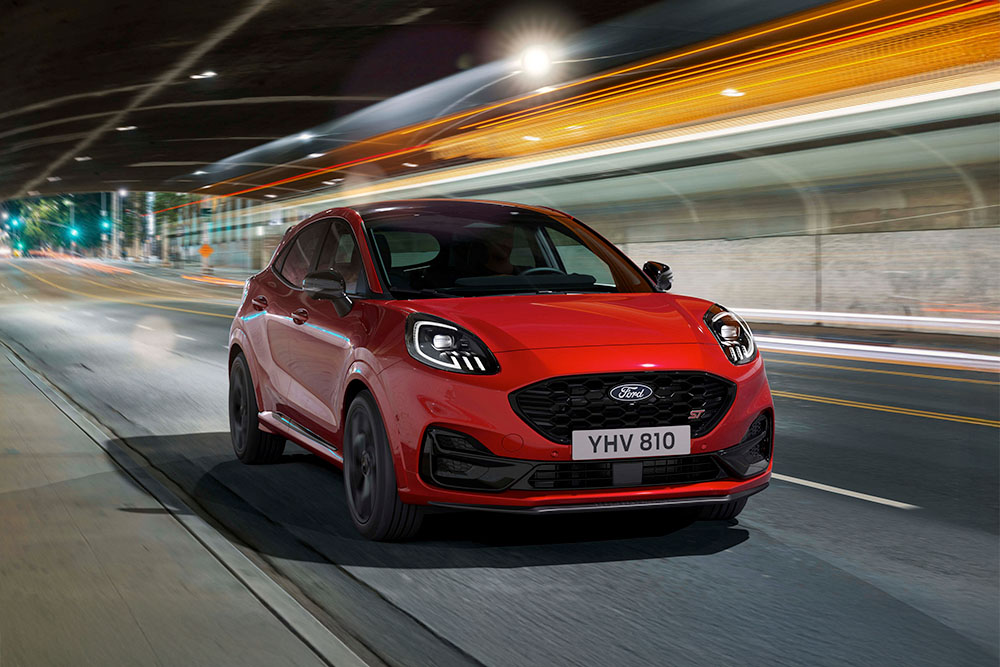 Een rode, sportieve Ford Puma ST-SUV die 's nachts over een helder verlichte stadsweg rijdt.
