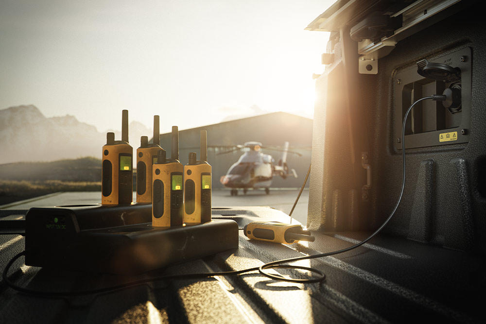 Meerdere walkie-talkies die worden opgeladen op de laadvloer van een Ranger Raptor bij zonsondergang, met een helikopter op de achtergrond.