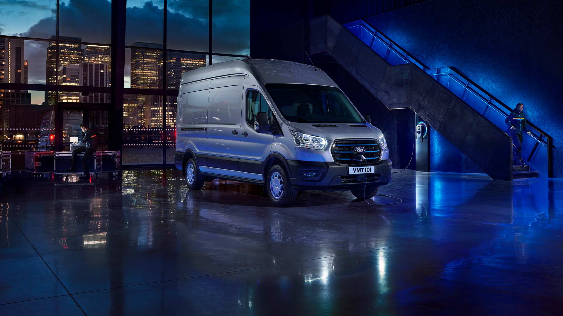Zilveren Ford E-Transit elektrische bestelwagen die binnen staat geparkeerd, met door de grote ramen een uitzicht op de skyline van een stad. 
