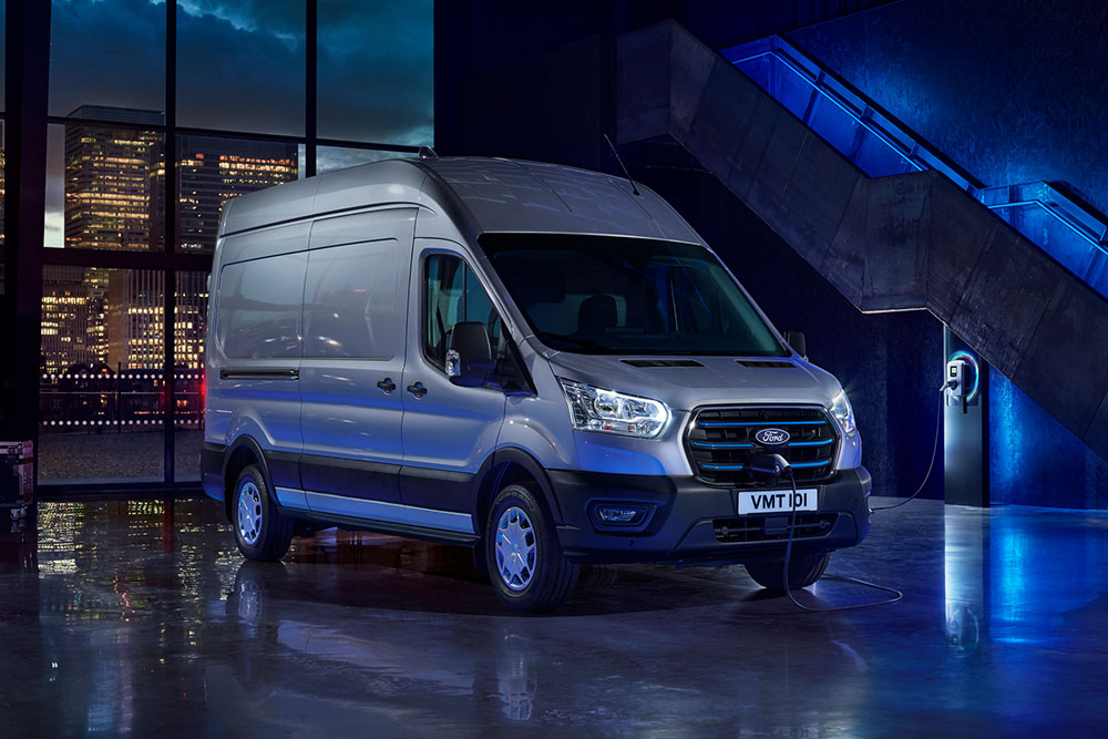 Zilveren Ford E-Transit elektrische bestelwagen die binnen staat geparkeerd, met door de grote ramen een uitzicht op de skyline van een stad. 