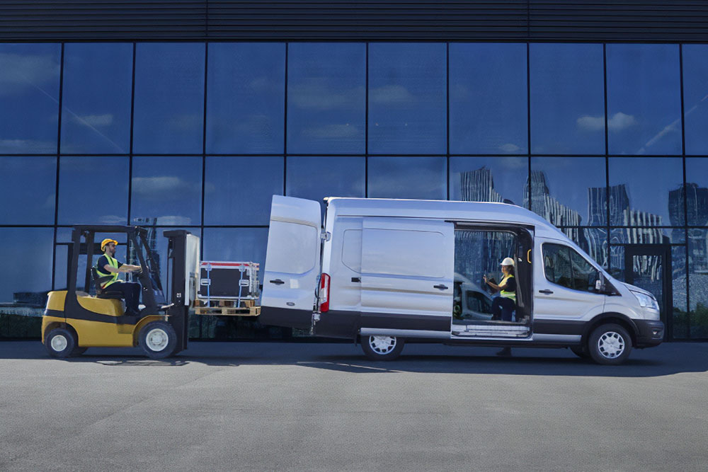 Twee mensen laden met een vorkheftruck spullen in een zilveren Ford Transit-bestelwagen.