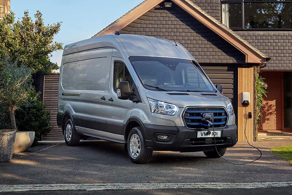 Zilveren Ford E-Transit-bestelwagen geparkeerd in een oprit naast een huis, aangesloten op een laadstation.
