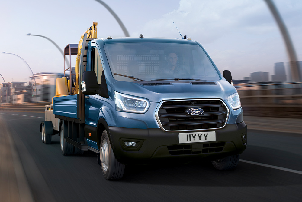 Vooraanzicht van de grille van een Ford Transit-chassis met cabine met een graafmachine die langs een stadsweg rijdt. 