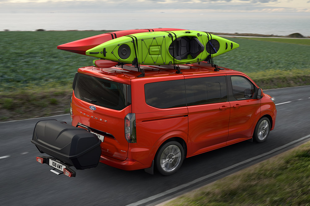 Rode Ford Pro Transit met twee kajaks op het dak, die een bagagedrager trekt en langs een kustweg rijdt.