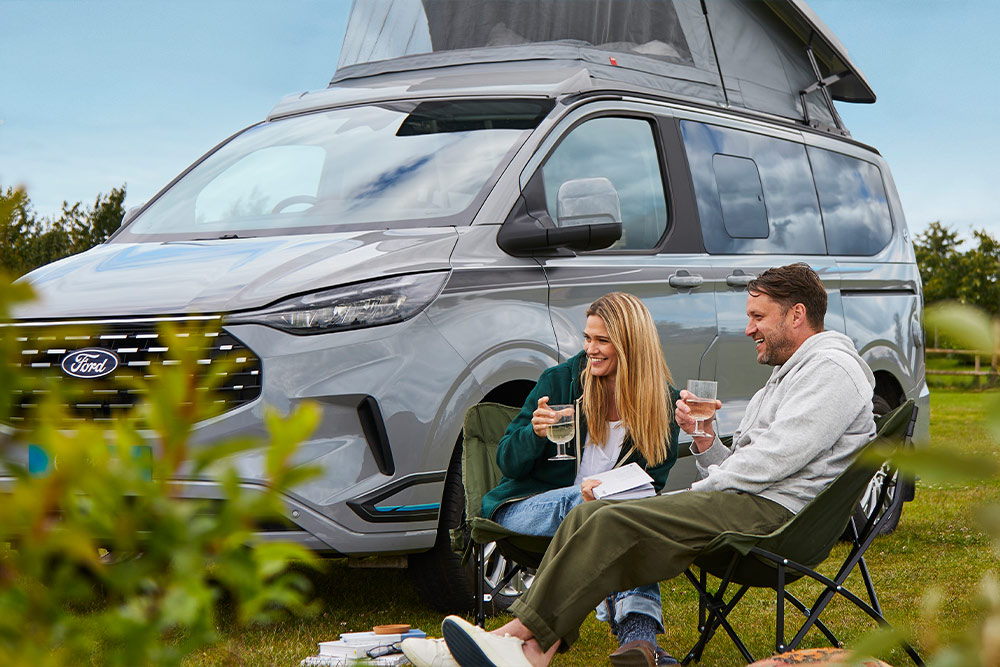Een stel geniet van drankjes in campingstoelen naast een grijze Ford Transit Custom campervan met een hefdak.