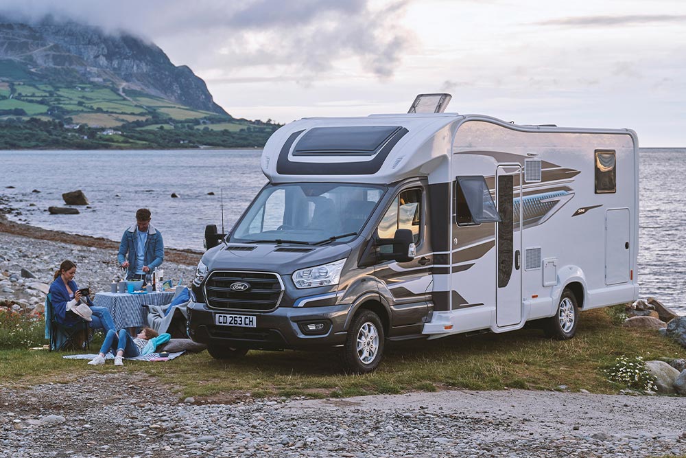 Een stel geniet van drankjes in campingstoelen naast een grijze Ford Transit Custom campervan met een hefdak.