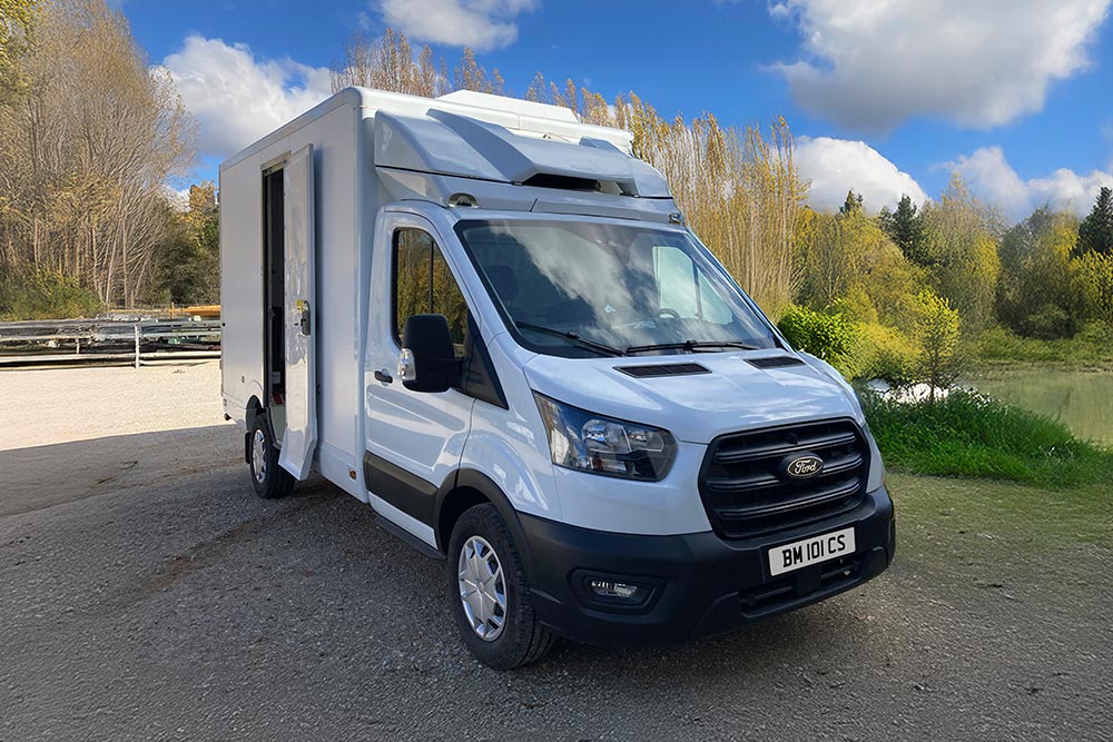 Vooraanzicht 3/4 van een witte Ford Transit bestelwagen geparkeerd bij een meer met bomen.