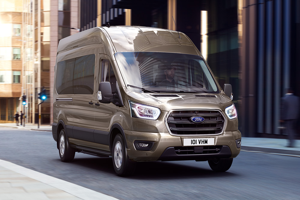 Vooraanzicht van een bronzen Ford Transit bestelwagen rijdend op een stadsstraat.