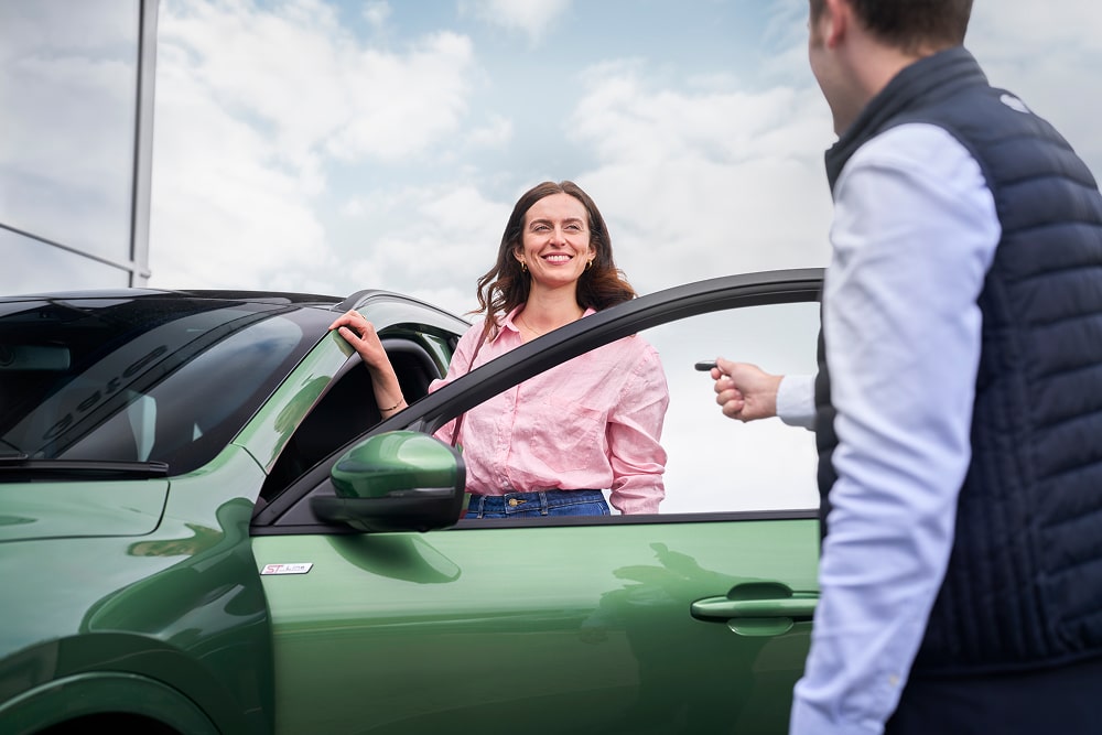 Een vrouw in een roze overhemd glimlacht naast een groene auto, terwijl een Ford-dealer haar de autosleutels overhandigt.