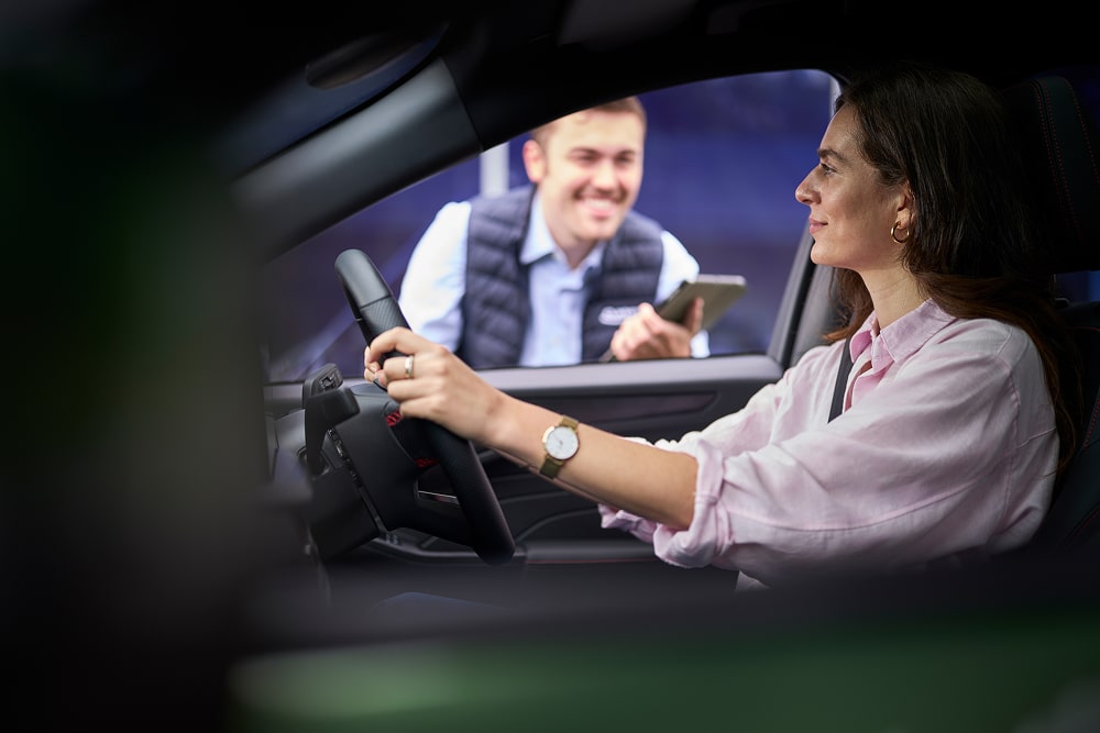 Een vrouw in een geparkeerde groene auto glimlacht naar een Ford-dealer achter het raam.